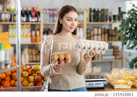 Young beautiful woman shopper in casual clothes chooses package of eggs in supermarket Young beautiful woman shopper in casual clothes chooses package of eggs in supermarket 135428861