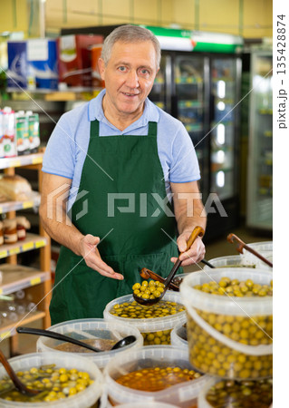 Portrait of smiling elderly salesman in apron arranging various types of pickled olives in hypermarket 135428874