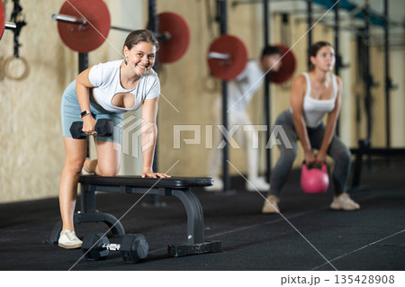 Athletic young woman pumping her biceps with a dumbbell while leaning on bench Athletic young woman pumping her biceps with a dumbbell while leaning on bench 135428908
