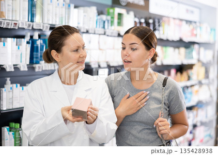 Female pharmacist advises a woman visitor a good skin care remedy, standing in the sales hall of pharmacy Female pharmacist advises a woman visitor a good skin care remedy, standing in the sales hall of pharmacy 135428938