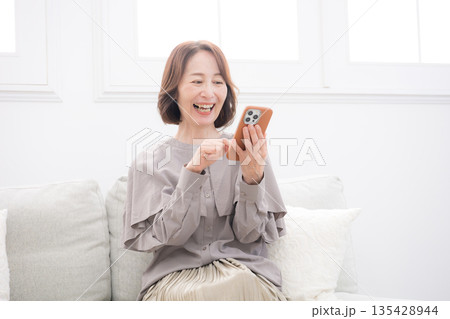 ソファに座りスマホを操作して予約や問い合わせをする綺麗な年配女性 135428944
