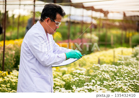Agricultural scientist in lab coat and gloves writing notes on clipboard while observing blooming flowers in a greenhouse, symbolizing plant research, floriculture, and sustainable agriculture 135432410