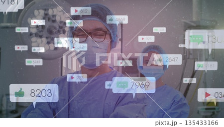Male surgeon and scrub nurse reviewing monitors under surgical light in OR, with social icons 135433166