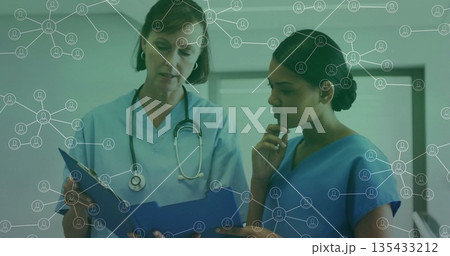 Nurses wearing scrubs reviewing patient records in clinic corridor, using blue folder, stethoscope 135433212