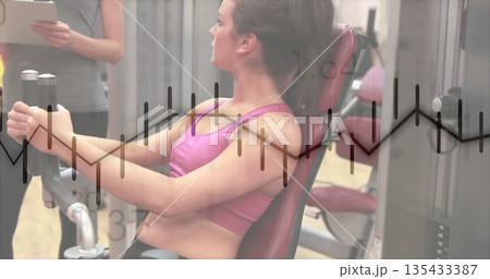 Woman in pink sports bra and leggings pushing chest-press handles in gym, with trainer and tablet Woman in pink sports bra and leggings pushing chest-press handles in gym, with trainer and tablet 135433387