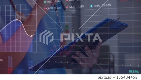 Doctor in blue scrubs and stethoscope holding clipboard in hospital with financial graphs overlay Doctor in blue scrubs and stethoscope holding clipboard in hospital with financial graphs overlay 135434458
