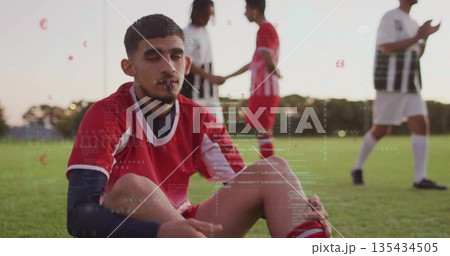 Sitting male soccer player drawing knees up and wearing red jersey on grass field, with cleats Sitting male soccer player drawing knees up and wearing red jersey on grass field, with cleats 135434505
