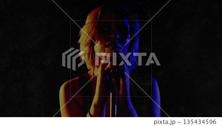 Performing teenage female singer gripping handheld microphone on dark stage, with red blue lighting 135434596