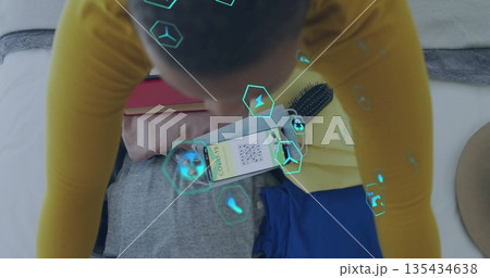 Woman in mustard top kneeling in bedroom scanning QR code on smartphone with clothes, teal icons 135434638