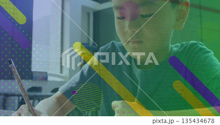 Writing boy in teal shirt sitting at home desk under blinds, using red-and-white pencil on paper 135434678
