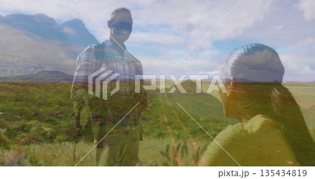 Standing man wearing plaid shirt sunglasses backpack facing kneeling partner in field, dirt trail Standing man wearing plaid shirt sunglasses backpack facing kneeling partner in field, dirt trail 135434819