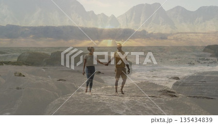 Holding hands couple walking at sandy shoreline with rocks, ocean waves and mountain range 135434839
