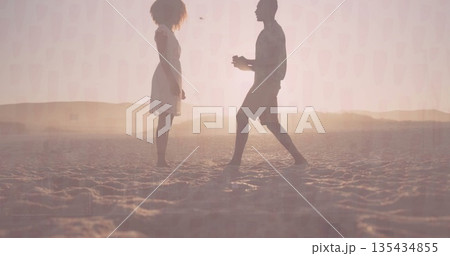Offering small box, man moving toward woman on rippled sandy shore at sunset, dune silhouettes 135434855