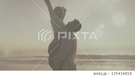 Lifting Hispanic man hoisting White woman high at sandy shoreline, with ocean waves and lens flare 135434858