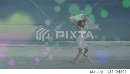 Woman in white dress running along beach holding white scarf, with ocean waves and bokeh overlays 135434863