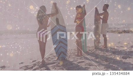 Standing friends in swimwear holding surfboards at sandy shoreline, with bright sunlight and waves 135434884