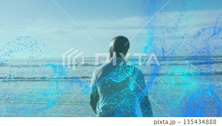 Standing man in grey shirt gazing toward horizon on wet sand, with waves, digital blue overlay 135434888