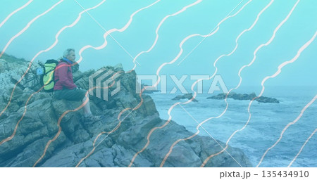Sitting hiker surveying choppy sea at coastal cliff, with yellow backpack, water bottle, copy space 135434910