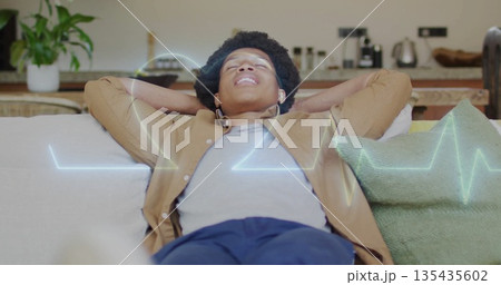 Reclining man holding hands behind head on living room sofa, with wireless earbuds and waveform 135435602