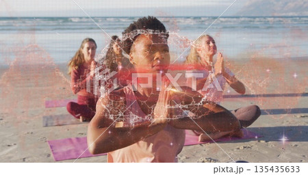Meditating adult woman in pink sleeveless top holding prayer pose on sandy beach, with yoga mats 135435633