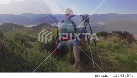 Resting mature hiker raising hand to face on grassy hillside, with red jacket, poles and backpack 135435673