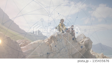 Sitting hiking partners viewing translucent network overlay on coastal rock outcrop, backpack Sitting hiking partners viewing translucent network overlay on coastal rock outcrop, backpack 135435676