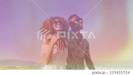 Smiling couple standing on sandy shoreline, wearing sunglasses with white dress and tropical shirt 135435724