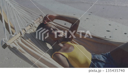 Reclining woman wearing yellow tank top shielding eyes from sun on sandy beach hammock, with waves 135435740