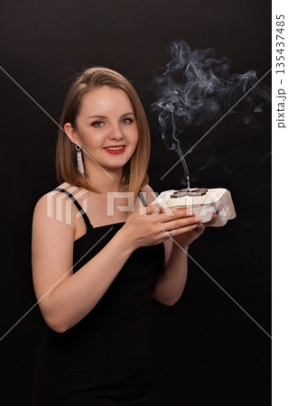 A woman in a black dress blows out a candle on a bento cake. Smoke on a black background. A girl's birthday, happiness, a gift. High quality photo 135437485