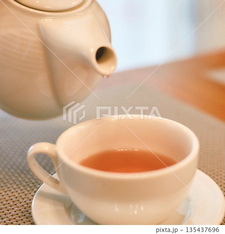 Warm cup of tea being poured from white teapot into a cup on a saucer in a cozy 135437696
