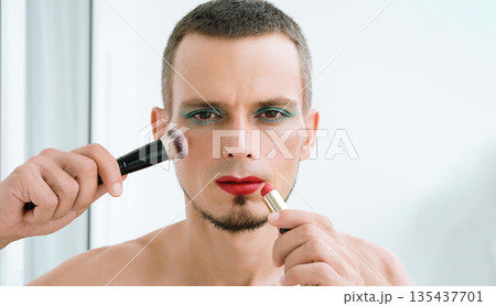 gay man with makeup and cosmetics on face. transgender man with bright eyeshadows paints lips with red lipstick and applies a blush brush to his face on white background 135437701