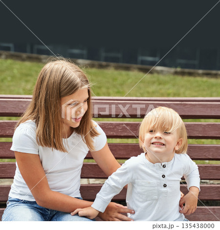 Young caucasian female teen and child brother smiling on bench in park on sunny 135438000