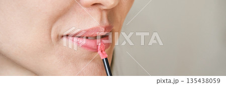 Caucasian female applying pink lipstick close-up with lip gloss brush 135438059