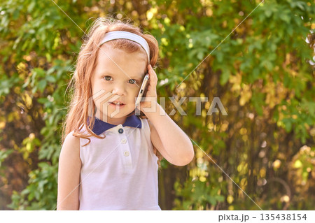 Young caucasian female child with red hair talking on phone outdoors in park 135438154