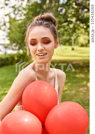 Young caucasian female with red balloons smiling outdoors in sunny park setting 135438169