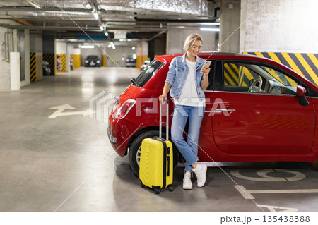 A woman stands in a parking garage with her luggage, looking at her phone next to a red car A woman stands in a parking garage with her luggage, looking at her phone next to a red car 135438388