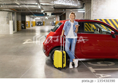 A woman stands in a parking garage with her luggage, looking at her phone near a red car 135438390