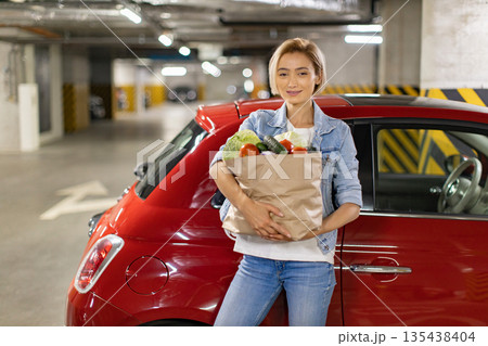 A woman smiles while holding a bag of fresh groceries next to her red car in a parking garage 135438404