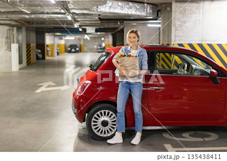 A woman smiles while holding a bag of groceries next to her red car in a parking garage A woman smiles while holding a bag of groceries next to her red car in a parking garage 135438411