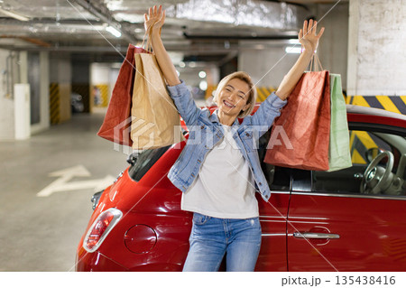 A happy woman stands next to her red car in a parking garage, holding shopping bags 135438416