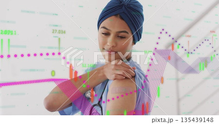 Touching bandage on arm, nurse in scrubs and turban in studio with stethoscope and financial chart 135439148