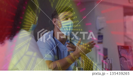 Gesturing doctor in scrubs and mask, speaking in consultation room with laptop and digital overlay 135439265
