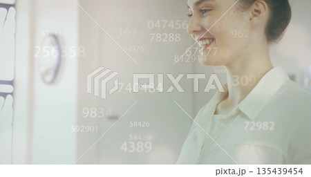 Smiling professional woman viewing floating numeric overlays in office workspace, with wall clock 135439454