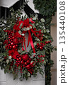 Festive red and green Christmas wreath with ornaments and ribbons displayed on a wall 135440108