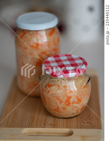 Jars of fermented sauerkraut with carrots on wooden cutting board in kitchen setting 135440112