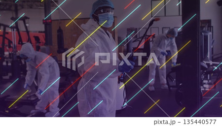 Sanitizing worker wearing protective suit holding sprayer wand cleaning treadmill at gym 135440577