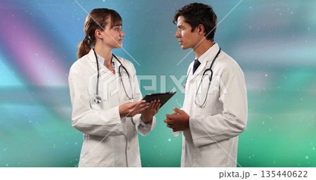 Two doctors wearing white coats holding tablet and discussing in studio, with two stethoscopes Two doctors wearing white coats holding tablet and discussing in studio, with two stethoscopes 135440622