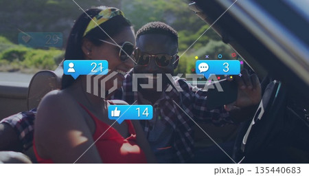 Taking selfie sunglasses-clad couple in convertible on scenic road, with phone and overlay icons 135440683