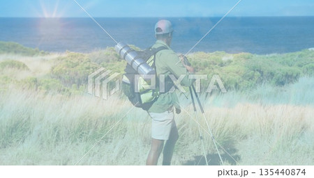 Standing hiker wearing cap placing tripod on dune overlooking sea with hiking backpack, copy space 135440874
