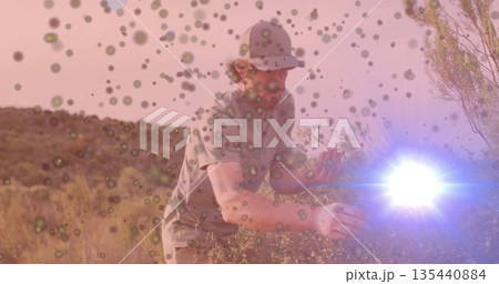 Crouching male wearing baseball cap, t-shirt examining shrubs on hillside, with lens flare overlays 135440884